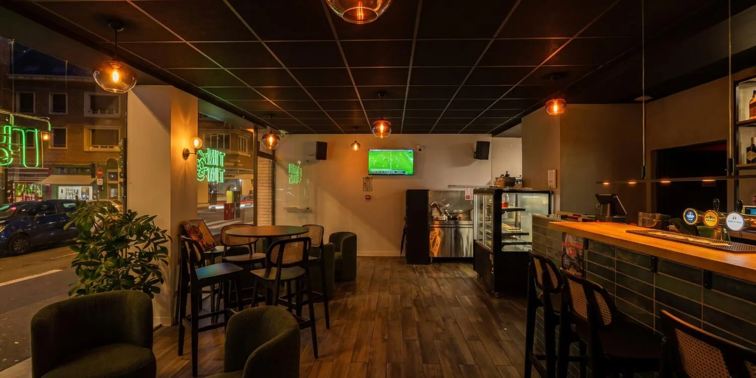 Salle côté rue, tables hautes, fauteuils verts et néon en vitrine.